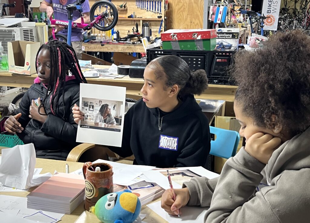 three Black girls sitting at a table journaling and discussing a photograph of a woman.