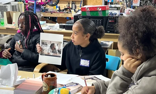 three Black girls sitting at a table journaling and discussing a photograph of a woman.